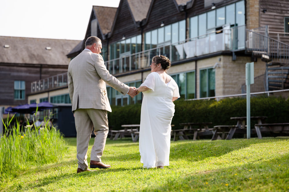 Newlyweds walking hand in hand on the lawn at De Vere Cotswold Water Park — hotel glass frontage behind them