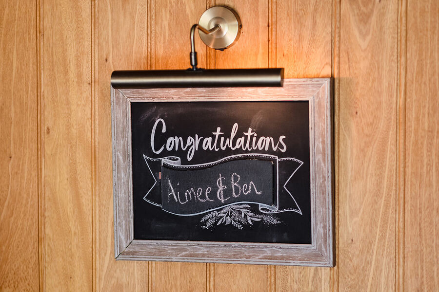Congratulations chalkboard sign with Curradine Barns wedding couple's names, lit by a brass picture light