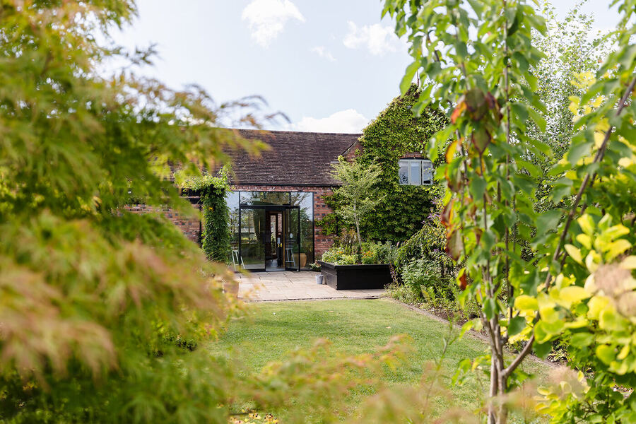 Curradine Barns gardens viewed through trees, with the red brick barn and glass extension beyond
