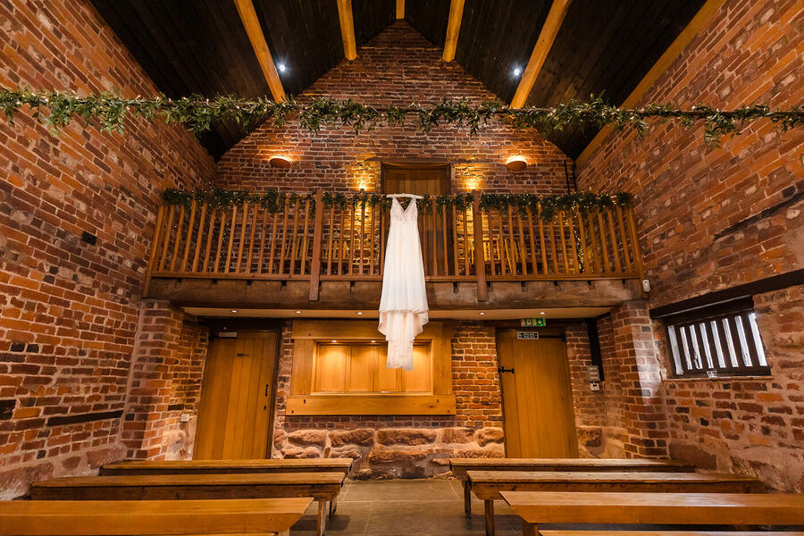 The Granary Barn ceremony room at Curradine Barns, Worcestershire — exposed red brick walls, oak beams and minstrel gallery with wedding dress hanging overhead